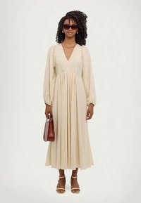 Nainen, jolla on kiharat hiukset, käyttää aurinkolaseja, pitkää kermaista mekkoa, jossa on pitkät hihat, keltaisia sandaaleja ja pitää ruskeaa käsilaukkua.