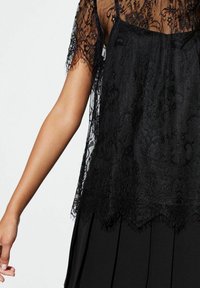 Black lace top with short sleeves, layered over a black pleated skirt, featuring intricate floral patterns and a scalloped hem detail.