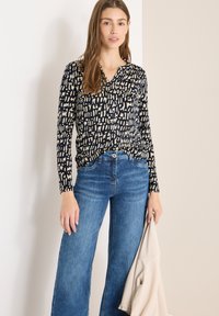 Marineblå bluse med cremefarvet abstrakt mønster i knap-til-ned-stil; kombineret med lyseblå bukser med vide ben. Modellen holder en beige jakke.