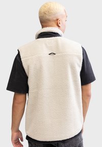 Gilet en polaire couleur crème avec une surface texturée, design sans manches et un petit détail noir à l'arrière, porté sur un t-shirt noir.