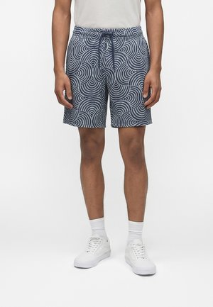Homme portant un short bleu marine et gris avec des motifs de cercles concentriques, des chaussettes blanches, des baskets blanches et un t-shirt blanc uni.