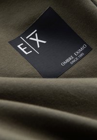 Olivegrüner Stoff mit einem schwarzen Etikett, auf dem die Buchstaben "E/X" und der Text "OMBRE EXIMIO SINCE 1985" zu sehen sind, mit einer glatten Textur.