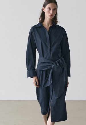 Donna che indossa un vestito blu navy a maniche lunghe con bottoni e una grande cintura annodata in vita, in piedi contro uno sfondo grigio chiaro uniforme.