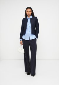 Vrouw in een marineblauwe blazer, blauwe gestreepte blouse, wijde marineblauwe broek, zwarte hakken, met een kleine witte handtas, staande tegen een witte achtergrond.