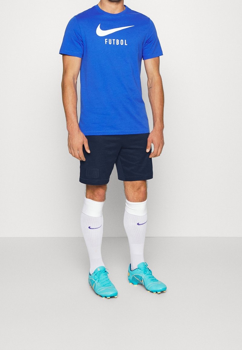 T-shirt bleu Nike avec logo blanc "FUTBOL", short bleu marine, chaussettes blanches avec accents violets, et chaussures de football turquoises.