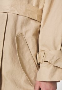 Manche de trench beige avec sangle ajustable et bouton près du poignet, et une poche latérale en biais avec des coutures visibles.