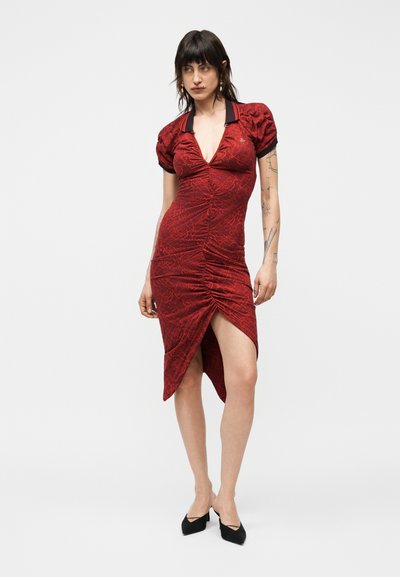 Γυναίκα που φοράει κόκκινο ασύμμετρο φόρεμα με ρυτιδωτό ύφασμα και κοντά φουσκωτά μανίκια, με μαύρα μυτερά τακούνια, στέκεται μπροστά σε απλό λευκό φόντο.