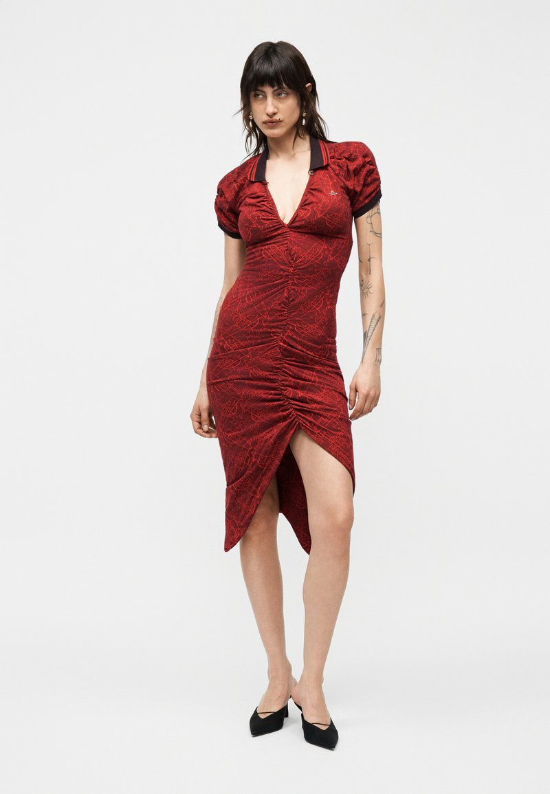 Woman wearing a red ruched asymmetrical dress with short puff sleeves and black pointed heels, standing against a plain white background.