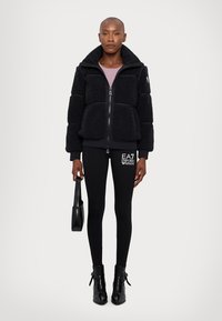 Svart fleecejacka med hög krage, dragkedjeslutning och mjuk textur; matchad med svarta leggings med logotypdetalj och ankelstövlar.