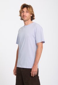 Homme aux cheveux blonds mi-longs, portant un T-shirt violet clair et un pantalon foncé, debout devant un fond blanc.
