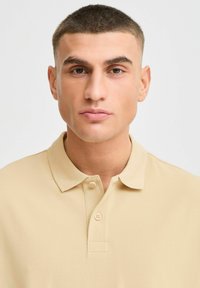 Beige polo skjorte med tre-knappers knappestolpe, klassisk krage og teksturert stofffinish. Nærbilde av overkroppen, nøytral bakgrunn.