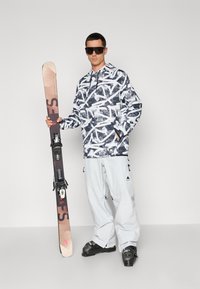 Snowboardista v vzorovanej čierno-bielej mikine s kapucňou, šedých lyžiarskych nohaviciach a v topánkach, ktorý drží hnedé lyže s viazaním na jednoduchom pozadí.