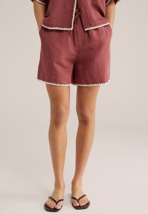 Osoba nosiaca hrdzavohnedé šortky a zodpovedajúci top s bielym lemom, stojaca bosá v čiernych žabkách na jednofarebnom pozadí.