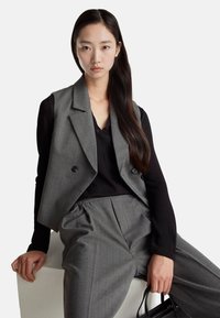 Szary kamizelka w prążki na miarę noszona na czarnej, długim rękawem bluzce, w połączeniu z szarymi spodniami. Modelka trzyma małą czarną torebkę, siedząc na białej powierzchni.