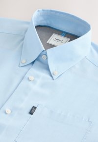 Lyseblå skjorte med knapper lavet af glat stof, med spids krave, enkelt brystlomme og et mærke på lommen.