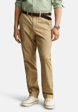 Man wearing beige pants, white sneakers, brown braided belt, green striped shirt, and a silver wristwatch, standing against a plain background.