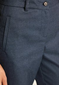 Närbild på marinblå randiga byxor med sidofickor och bruna knappar i midjan.