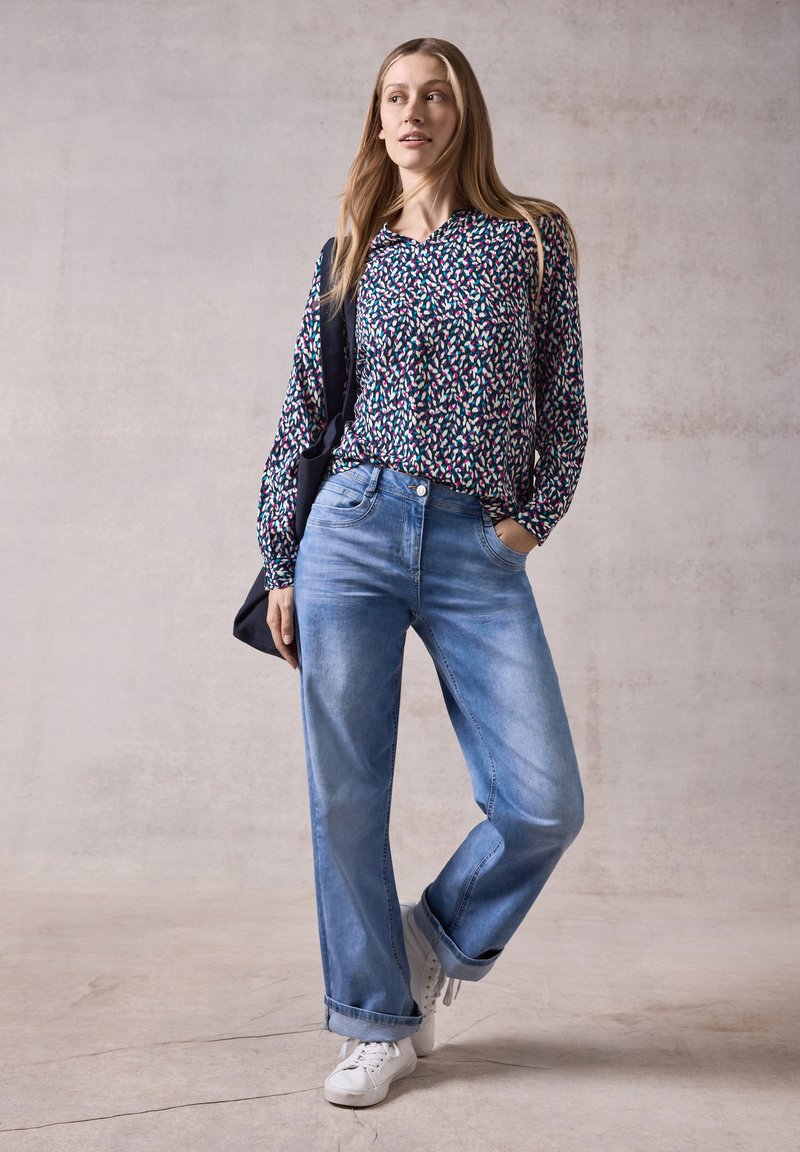 Un paio di jeans in denim azzurro chiaro con vestibilità rilassata, indossati con una camicia scura a fantasia. Sneakers bianche e una borsa a tracolla scura sono incluse.