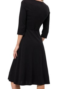 Femme portant une robe noire longueur genou avec manches trois-quarts, taille cintrée et jupe évasée, vue de dos.