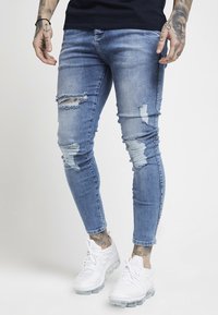 Jeans skinny desgastados de color azul claro con detalles rasgados y un ligero efecto de decoloración, combinados con zapatillas deportivas blancas y una camiseta negra.