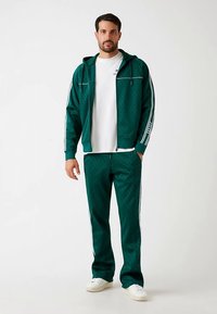 Chándal verde hecho de tela suave, con chaqueta con capucha y detalles en blanco, y pantalones con rayas laterales. Se lleva sobre una camiseta blanca.
