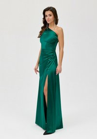 Smaragdově zelená saténová večerní šaty na jedno rameno, s řaseným designem, rozparkem do stehna a upnutým pasem, v kombinaci s černými špičatými podpatky.