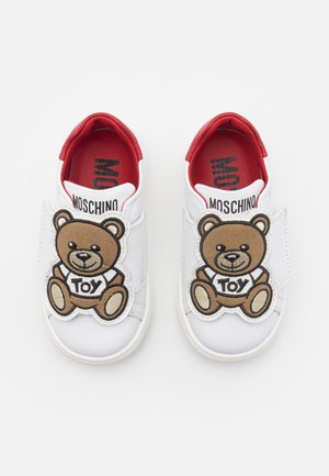 Hvide børnesneakers med rødt indre, hver med en stor brun bamsepatch mærket "TOY" på de forreste stropper.