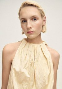 Beige sleeveless top with gathered fabric, featuring a halter neck design and soft texture, against a neutral background.