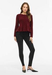 Bordowy sweter peplum z teksturowanej tkaniny, z okrągłym dekoltem i zapinany z przodu na guziki; w połączeniu z czarnymi obcisłymi jeansami i szpilkami w szpic.