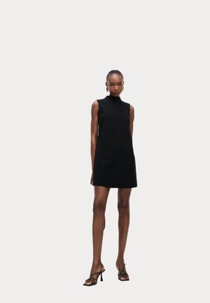 Schwarzes ärmelloses Kleid mit hohem Halsausschnitt, aus glattem Stoff gefertigt, mit weitem Schnitt und kurzem Saum, kombiniert mit Riemchenschuhen.