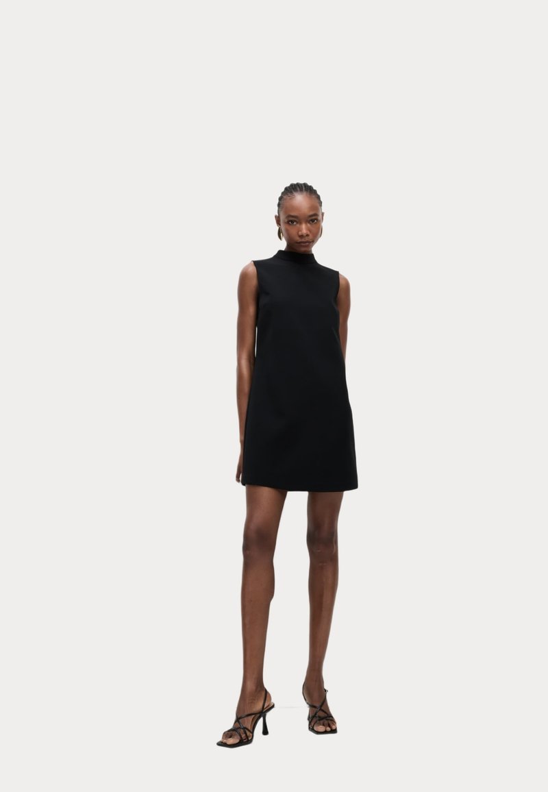 Schwarzes ärmelloses Kleid mit hohem Halsausschnitt, aus glattem Stoff gefertigt, mit weitem Schnitt und kurzem Saum, kombiniert mit Riemchenschuhen.