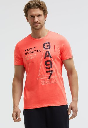 Mann trägt ein korallenfarbenes T-Shirt mit schwarz-weißen Yachtregatta- und Segelgrafiken, kombiniert mit dunklen Shorts, steht vor einem einfarbigen Hintergrund.