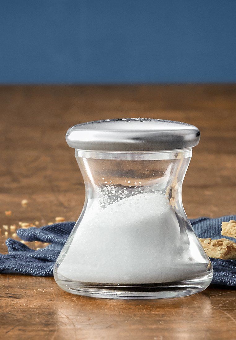 Duidelijke glazen zoutvaatje met een ronde vorm en een zilveren, ronde deksel. Bevat witte zout, geplaatst op een gestructureerde blauwe doek.