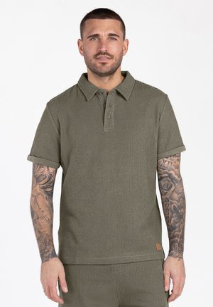 Man met kort haar en tatoeages op beide armen, gekleed in een olijfgroen, gestructureerd poloshirt met korte mouwen en bijpassende broek.