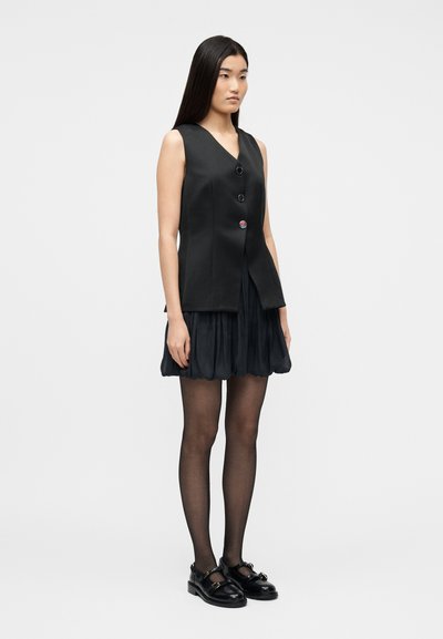 Γυναίκα που φοράει αμάνικο μαύρο γιλέκο με κουμπιά, κοντή μαύρη πλισέ φούστα, μαύρο διάφανο καλσόν και μαύρα παπούτσια Mary Jane, στέκεται σε λευκό φόντο.