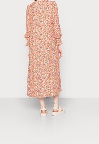 Robe longue à motifs avec des designs floraux vibrants et des couleurs orange, dotée de manches volantes. Portée avec des sandales à plateforme beiges.