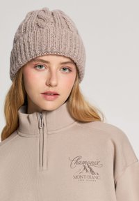 Jasnobeżowa, dziana czapka z teksturowanym wzorem, noszona z beżową bluzą z wysokim kołnierzem i zamkiem, na której znajduje się graficzny motyw górski oraz logo tekstowe.