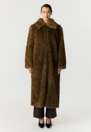 Une femme porte un long manteau de fourrure marron à col large, un pantalon foncé et des chaussures noires à talons, sur un fond blanc uni.