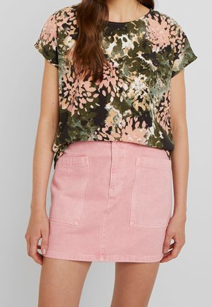 Vrouw die een kort mouw bloes met groene en roze bloemen draagt, ingestopt in een lichtroze denim minirok met steekzakken aan de voorkant.