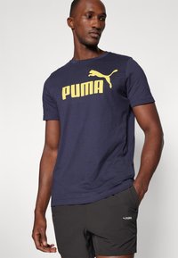 T-shirt en coton bleu marine avec un grand logo "PUMA" jaune et un graphique de puma en train de sauter. Associé à un short noir.