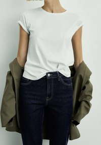 Camiseta de algodón blanca con mangas enrolladas, combinada con jeans de mezclilla oscura y un blazer verde oliva drapeado sobre los hombros.