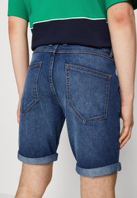 Persona vestida con pantalones cortos de mezclilla oscuros enrollados y una camisa en bloques de color verde y azul marino, mostrada desde la cintura hacia abajo por detrás.