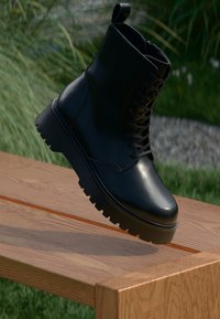 Juodas odinis kulkšnių botas su tekstūruota paviršiumi, raišteliais priekyje ir stambiu, grublėtu padu. Pateiktas ant medinės plokštės su žolės fonu.