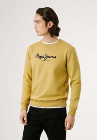 Felpa gialla in cotone con maniche lunghe, scollo rotondo e logo ricamato "Pepe Jeans London" in nero. Indossata sopra una camicia bianca.