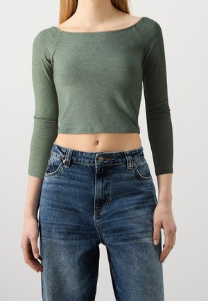 Personne portant un crop top vert moulant à manches longues et un jean denim bleu taille haute sur un fond uni.