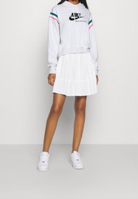 Sudadera gris con el logo negro de "Nike Sportswear", acentos a rayas multicolores en las mangas, combinada con una falda blanca con pliegues y zapatillas blancas.
