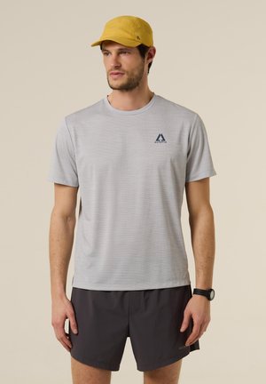 Man met gele pet, lichtgrijs shirt met korte mouwen en "Altavia"-logo, zwarte korte broek en zwarte polshorloge, staand tegen een beige achtergrond.