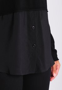 Svart lager-på-lager-skjorta med en texturerad övre del och en mjukare nedre sektion med fyra svarta knappar längs sidosen.