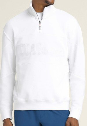 Uomo che indossa una felpa bianca con cerniera a quarto di giro e logo "Wilson" tono su tono sul petto, e pantaloni blu, che guarda leggermente a destra.