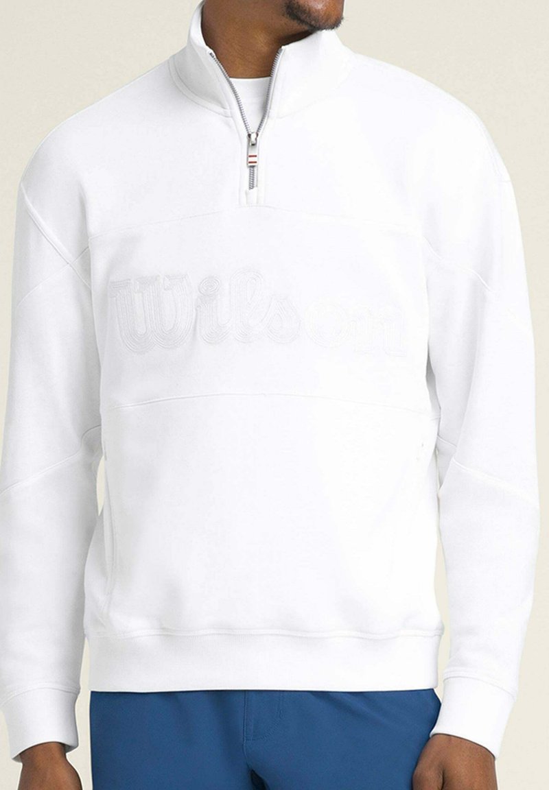 Homme portant un sweat-shirt blanc à fermeture éclair 1/4 avec le logo "Wilson" ton sur ton sur la poitrine et un pantalon bleu, légèrement tourné vers la droite.